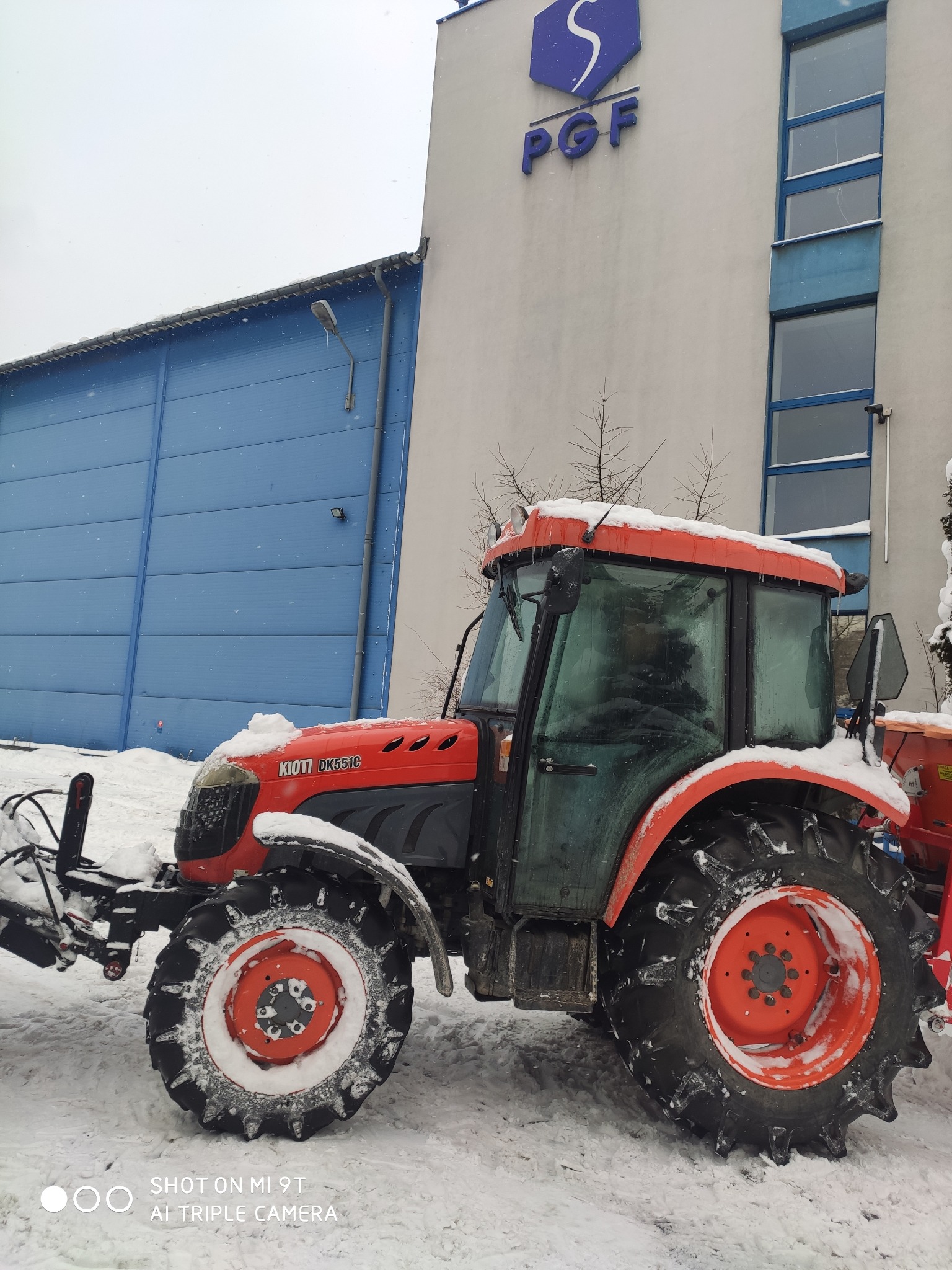 Czerwony traktor Kioti DK551C z pługiem do śniegu, częściowo pokryty śniegiem, stoi na tle błękitnego magazynu i budynku PGF w zimowy dzień.