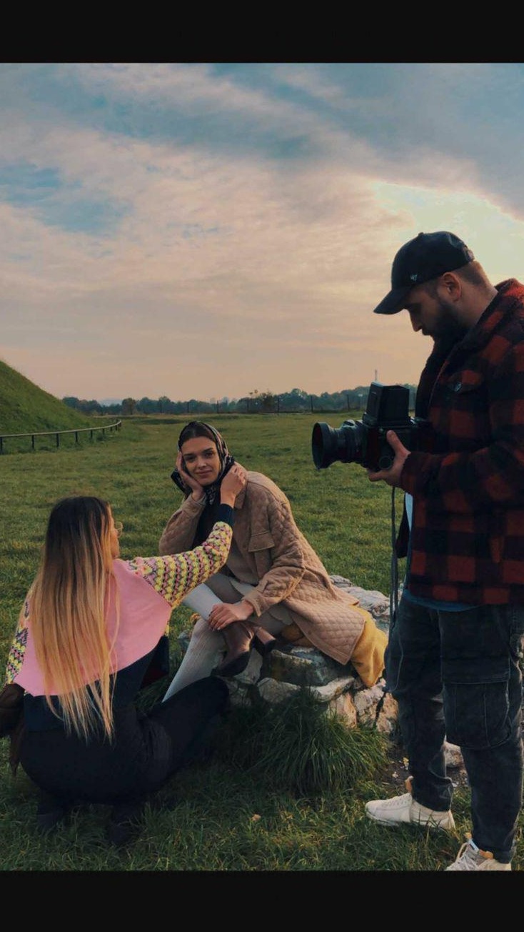 Sesja zdjęciowa w plenerze: modelka w chustce, beżowym płaszczu i jasnych rajstopach pozuje siedząc na kamieniu; stylistka poprawia chustkę modelki, a fotograf w czapce i czerwonej kraciastej...