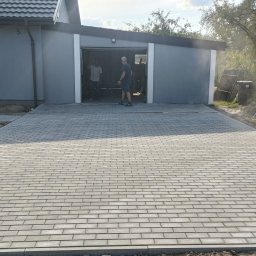 PHU Eurokop Justyna Zagaja - Pracownik bez koszulki zagęszcza nowo ułożoną, szarą kostkę brukową na podjeździe przed garażem, w tle widoczny budynek mieszkalny z brązowymi roletami.