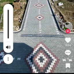 Długa, symetryczna ścieżka z kostki brukowej ułożona w geometryczne wzory, z widocznym odwodnieniem liniowym i paletami kostki w tle. Układanie kostki.