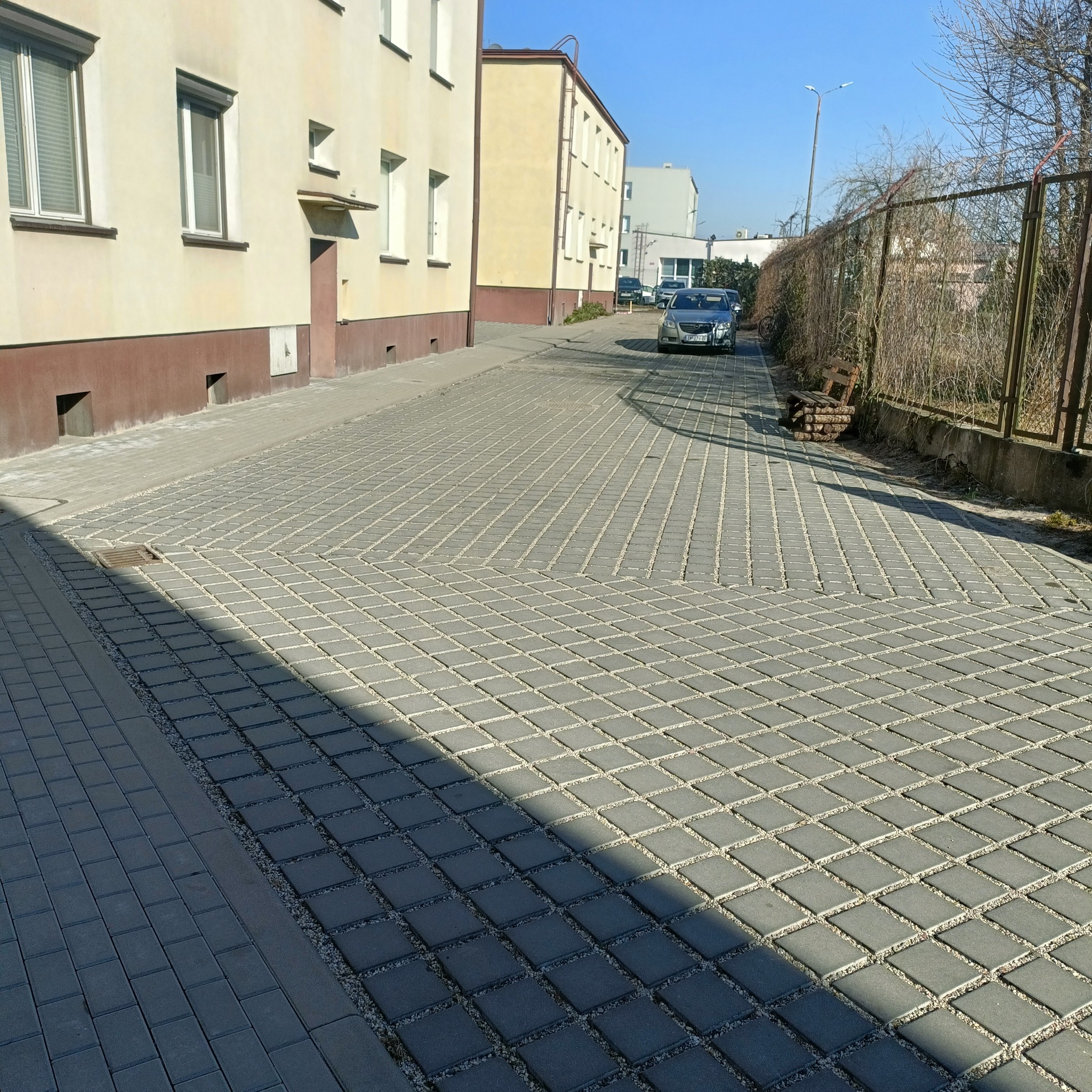 Szara kostka brukowa ułożona w dwa wzory: prostokątny i romboidalny, z jasnymi fugami wypełnionymi drobnym żwirem, cień drzewa na części powierzchni, budynek w tle.