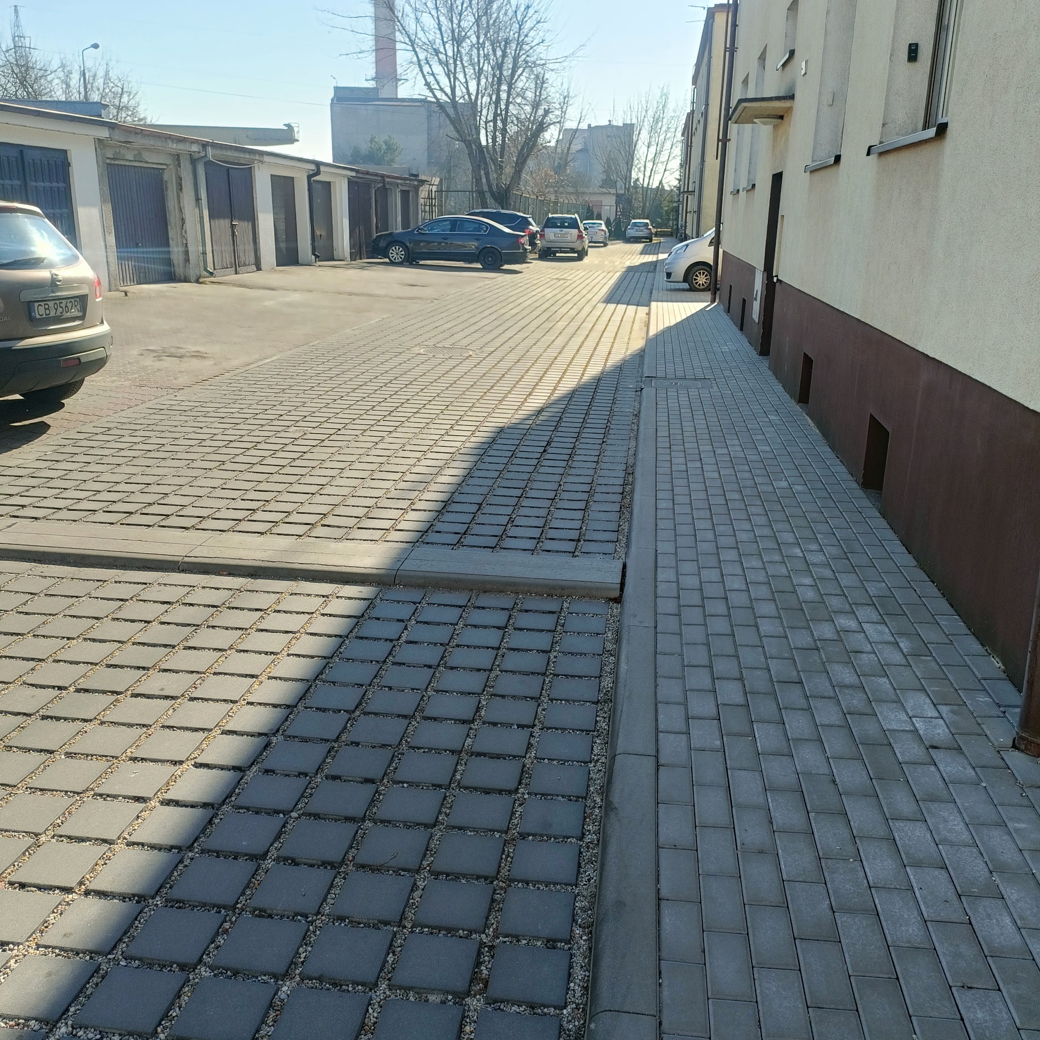 Ułożona szara kostka brukowa na parkingu z widocznymi garażami i zaparkowanymi samochodami w tle, z wyraźnym podziałem na dwa rodzaje kostki i odpływ liniowy.