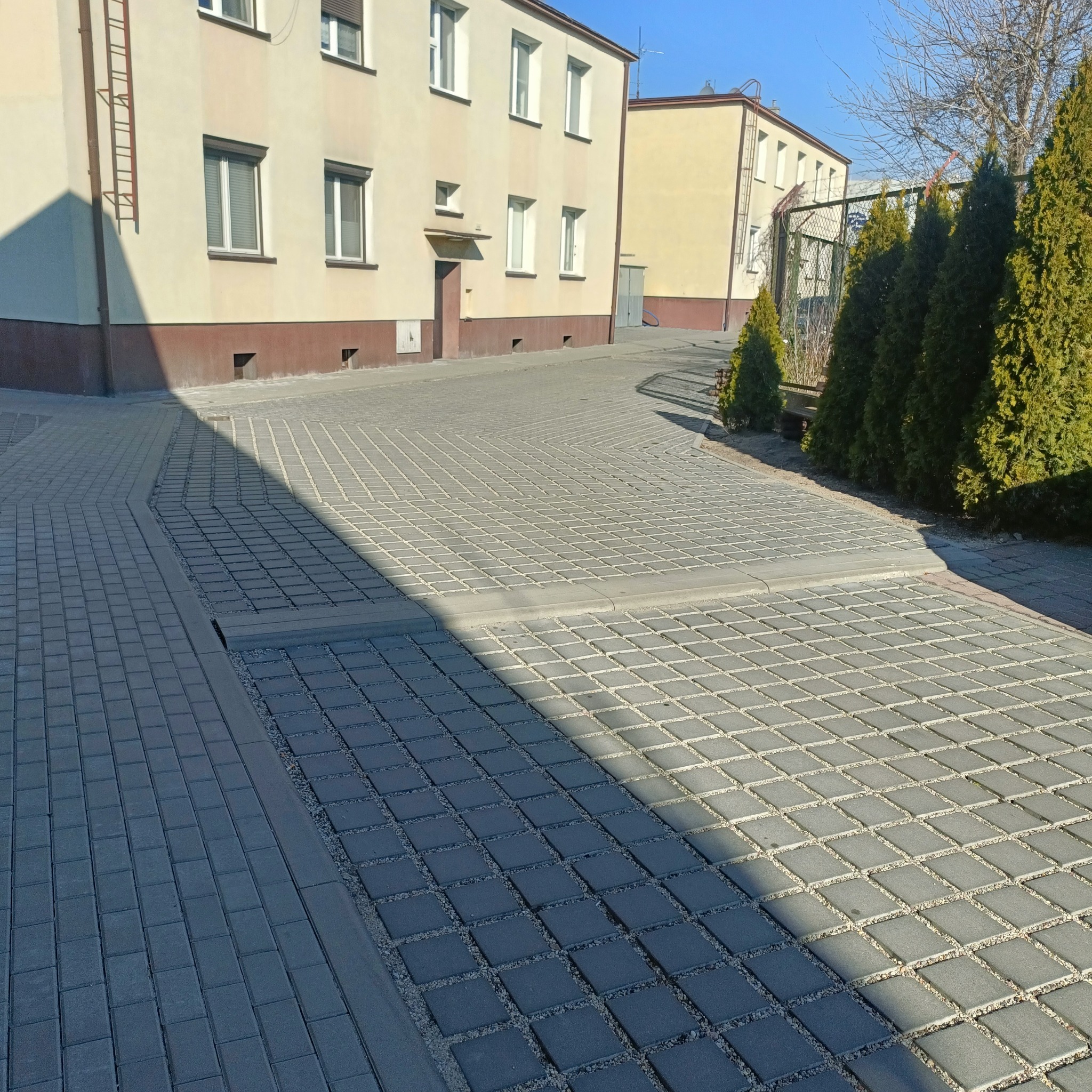Ułożona szara kostka ażurowa z wypełnieniem żwirowym na podjeździe przed budynkiem mieszkalnym, widoczne krawężniki i cień padający na powierzchnię.