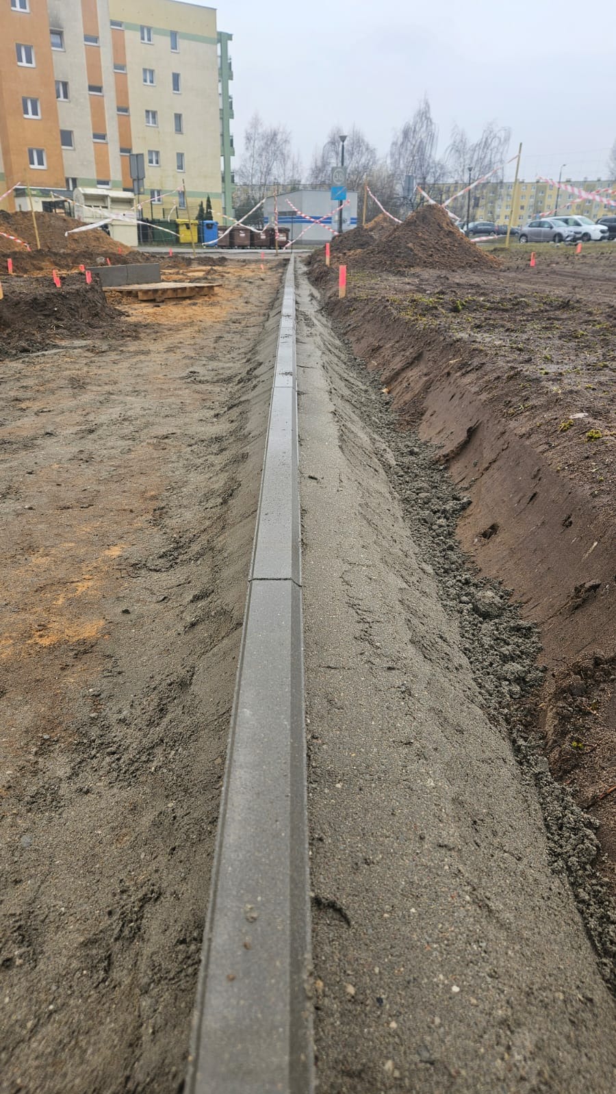 Świeżo ułożony obrzeżnik betonowy w gruncie, przygotowany do dalszych prac brukarskich, widoczne wykopane koryto i piasek podsypkowy, w tle budynek mieszkalny.