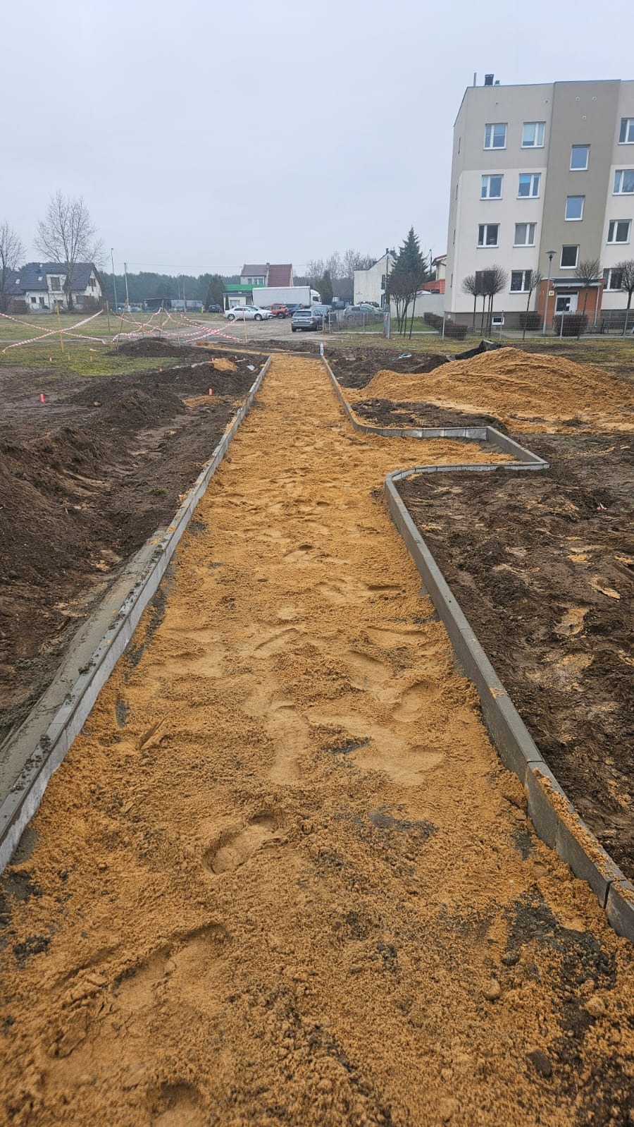 Przygotowany podkład piaskowy z obrzeżami betonowymi pod ścieżkę na terenie budowy, widoczne ślady stóp w piasku.
