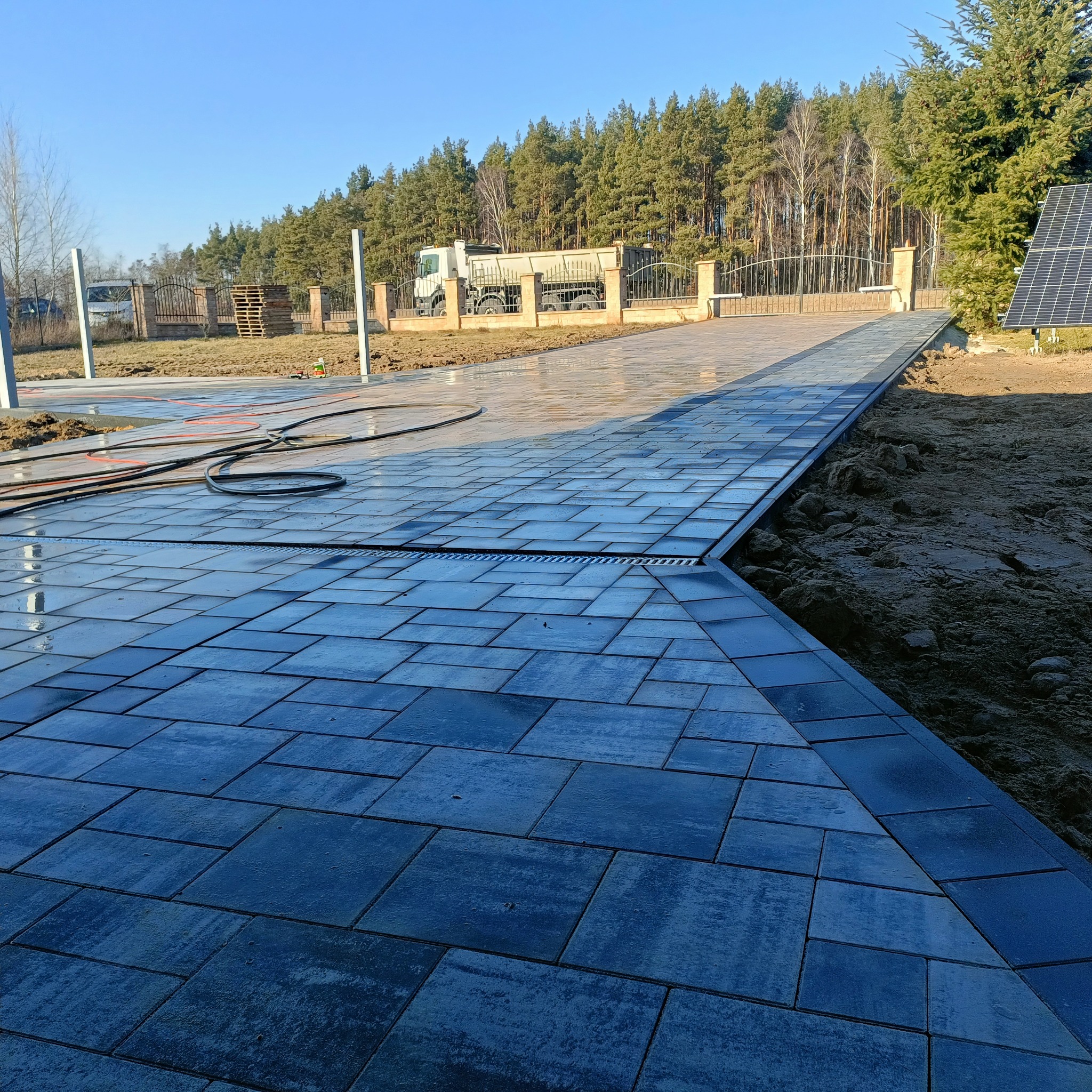 Świeżo ułożona, mokra, grafitowa kostka brukowa prowadząca do bramy wjazdowej, z widocznym odwodnieniem liniowym, zaparkowaną ciężarówką w tle i panelem słonecznym po prawej stronie.