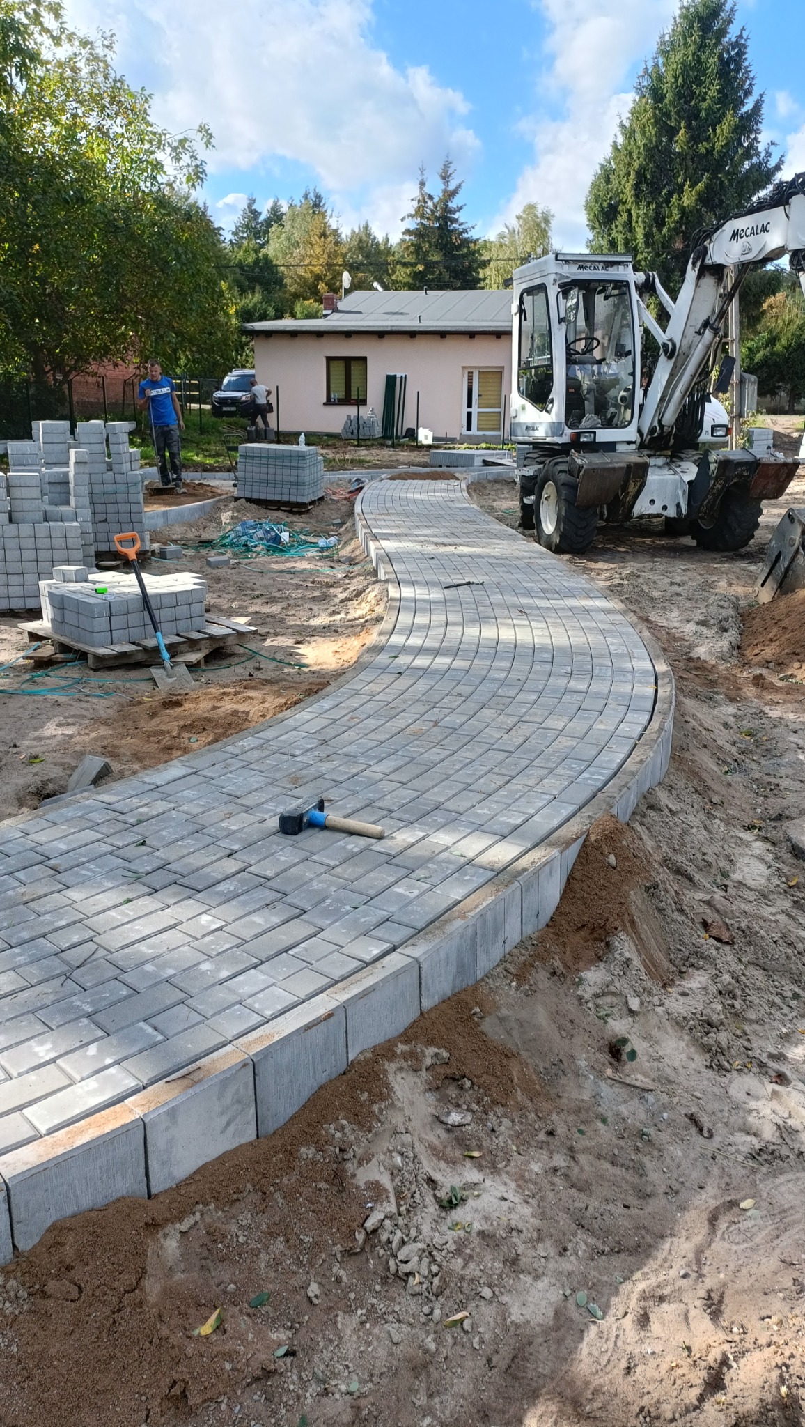 Układanie zakrzywionego chodnika z szarej kostki brukowej, obrzeża z betonowych krawężników, palety z kostką, koparka Mecalac w tle, robotnicy przy pracy.