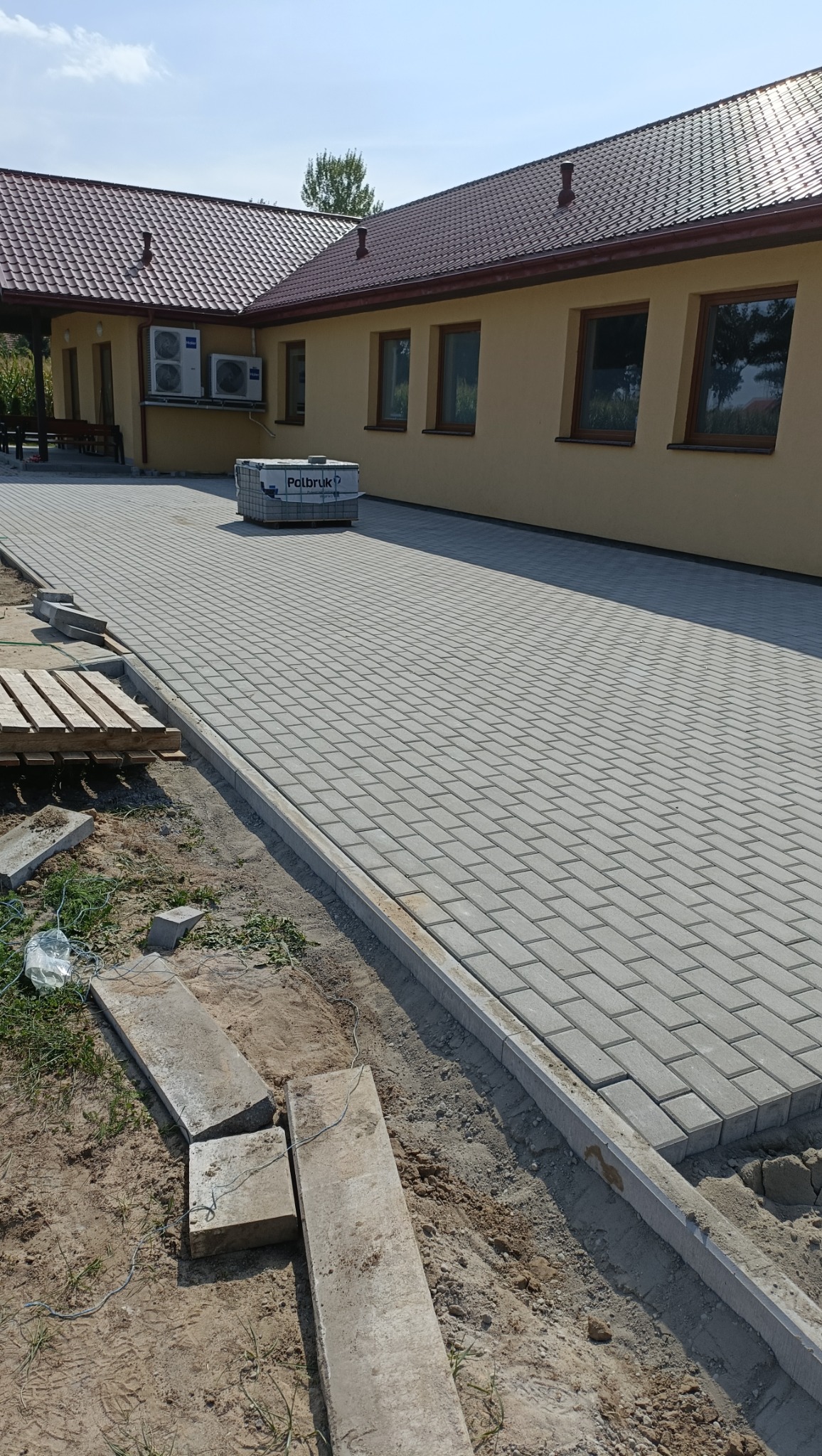 Świeżo ułożona, szara kostka brukowa na podjeździe, z paletą kostki w tle, obrzeża z betonu, widoczne niedociągnięcia w postaci piasku i resztek materiałów budowlanych.