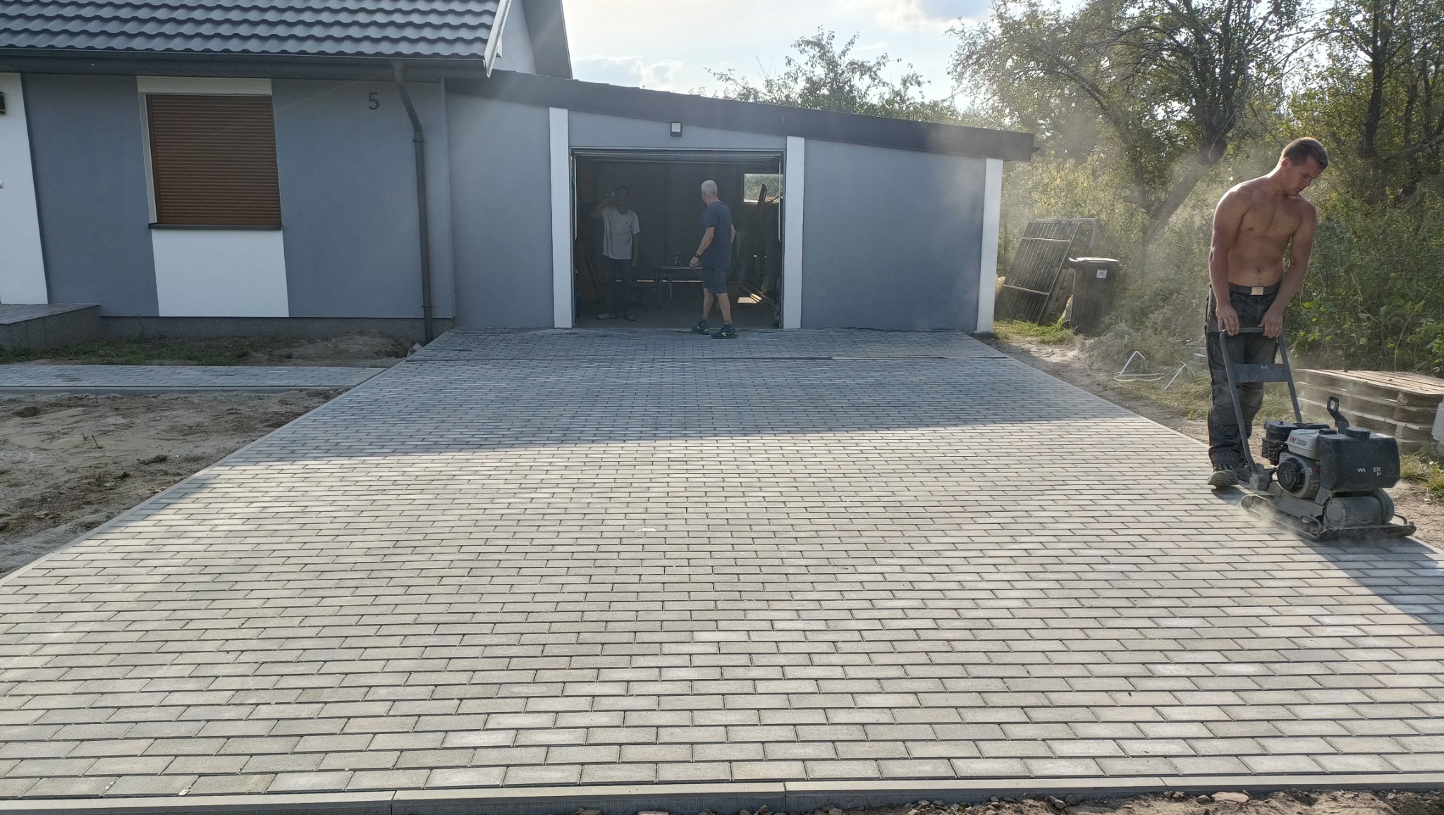 Pracownik bez koszulki zagęszcza nowo ułożoną, szarą kostkę brukową na podjeździe przed garażem, w tle widoczny budynek mieszkalny z brązowymi roletami.