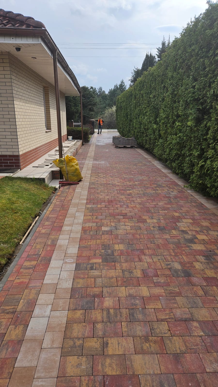 Kolorowa kostka brukowa tworząca ścieżkę obok domu z zielonym żywopłotem. W tle majster w pomarańczowej kamizelce. Widoczny worek i szczotka.