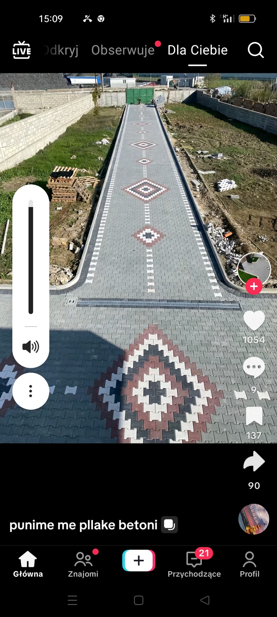 Długa, symetryczna ścieżka z kostki brukowej ułożona w geometryczne wzory, z widocznym odwodnieniem liniowym i paletami kostki w tle. Układanie kostki.