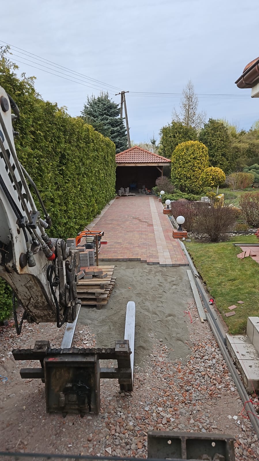 Układanie kostki: widok na częściowo wybrukowany podjazd z czerwono-beżowej kostki, palety z kostką i ubijarkę. W tle zielony żywopłot i zadaszona wiata.