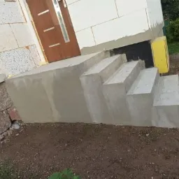 Nowo wykończone, surowe, betonowe schody wejściowe prowadzące do brązowych drzwi w budynku z ociepleniem styropianowym i fragmentem kamiennej ściany, widoczne z boku.