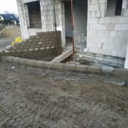 Budowa domu jednorodzinnego: fundamenty z bloczków betonowych, widoczny etap murowania ścian fundamentowych, teren wokół w stanie surowym.