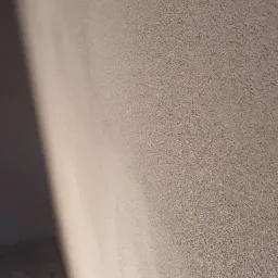 Świeżo nałożona, jasna, strukturalna powłoka gipsowa na ścianie w rogu pomieszczenia, z widocznym efektem cieniowania i surowym betonem na podłodze.