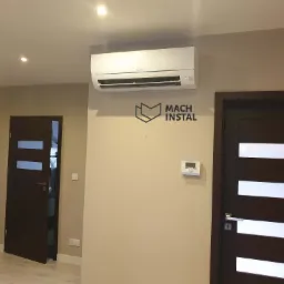 Biały klimatyzator Mitsubishi zamontowany na beżowej ścianie w pomieszczeniu z dwiema ciemnymi, nowoczesnymi drzwiami z przeszkleniami; pod klimatyzatorem widoczne logo firmy Mach Instal.