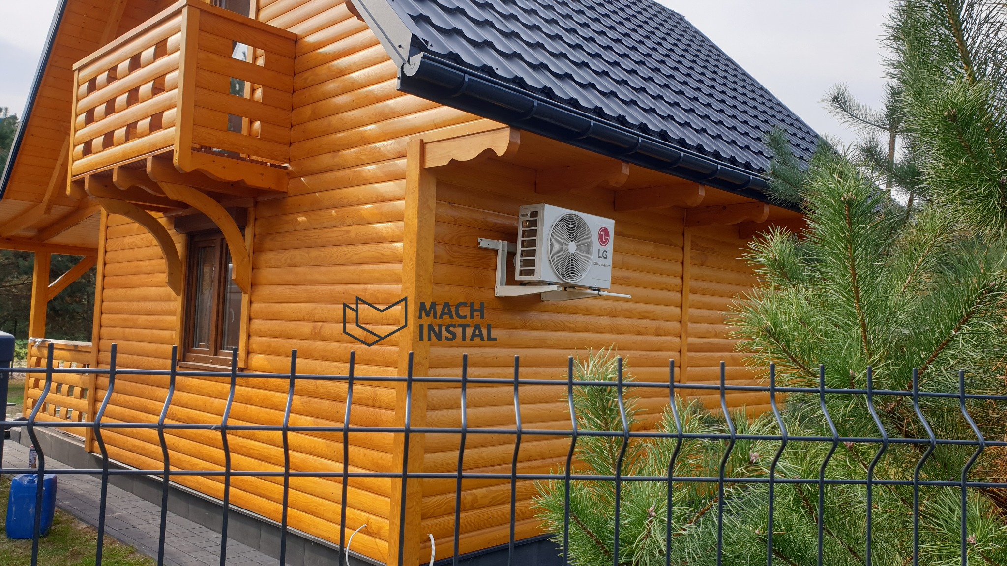 Zewnętrzna jednostka klimatyzacji LG zamontowana na drewnianej ścianie domu z bali, widoczna zza metalowego ogrodzenia i iglastego krzewu, w rogu widoczny niebieski kanister.