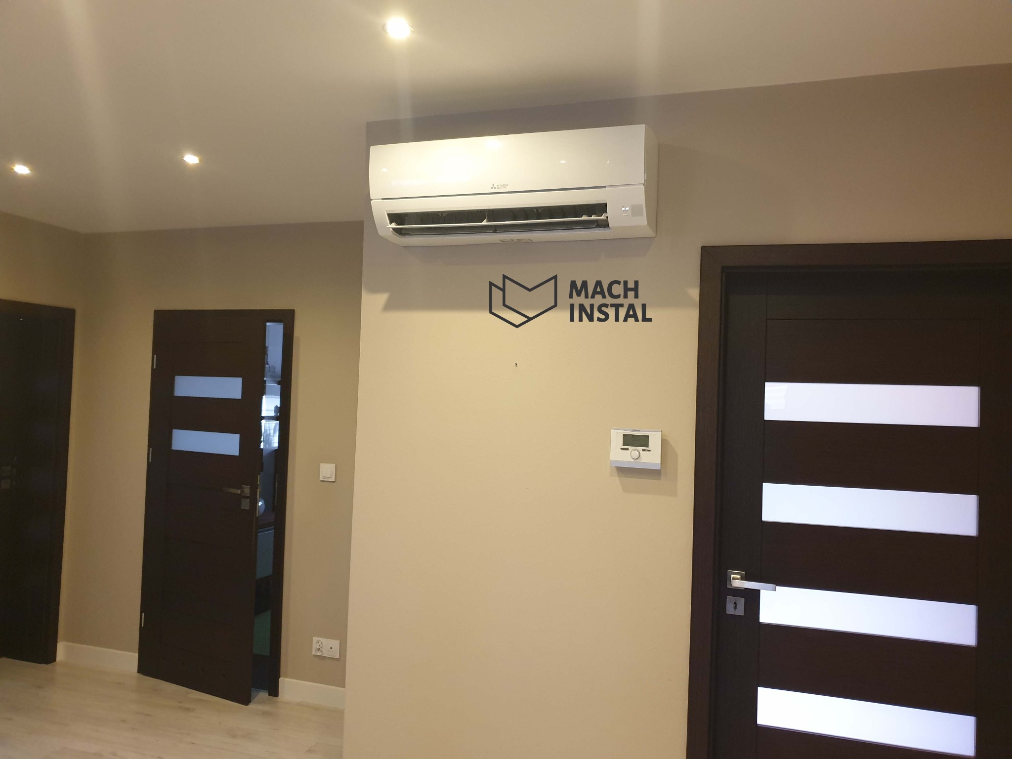 Biały klimatyzator Mitsubishi zamontowany na beżowej ścianie w pomieszczeniu z dwiema ciemnymi, nowoczesnymi drzwiami z przeszkleniami; pod klimatyzatorem widoczne logo firmy Mach Instal.