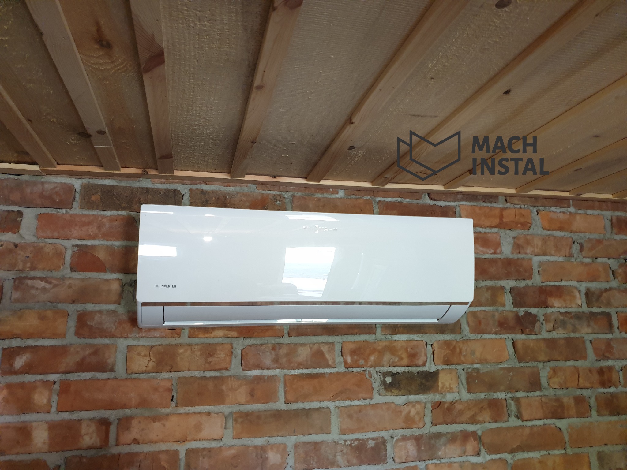 Biały klimatyzator Viessmann DC Inverter zamontowany na ceglanej ścianie z drewnianym sufitem. Widoczne logo firmy Mach Instal w prawym górnym rogu.