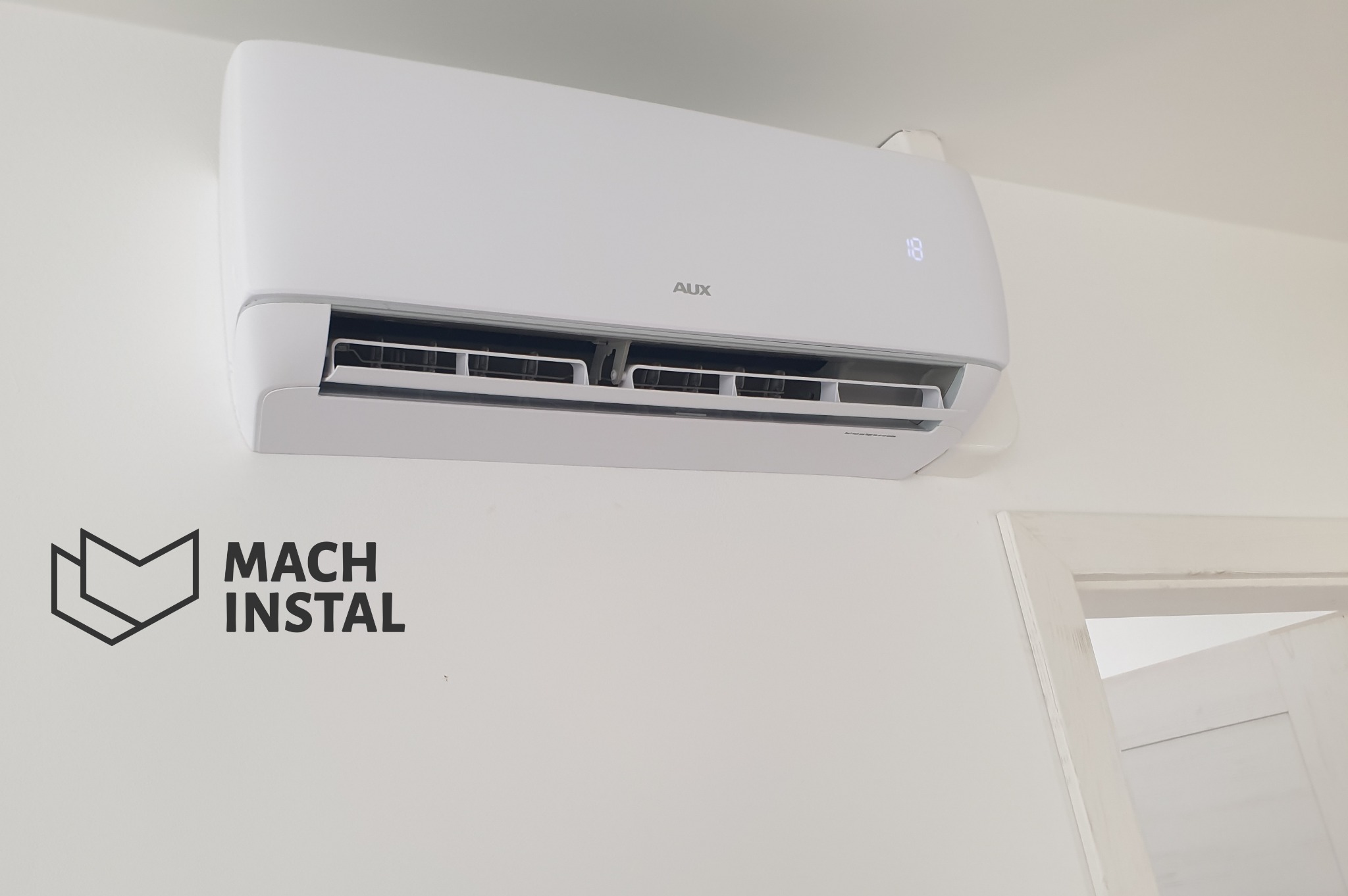 Biały klimatyzator AUX J-Smart zamontowany na białej ścianie, widoczne logo Mach Instal na ścianie poniżej urządzenia, fragment ościeżnicy drzwi w tle.
