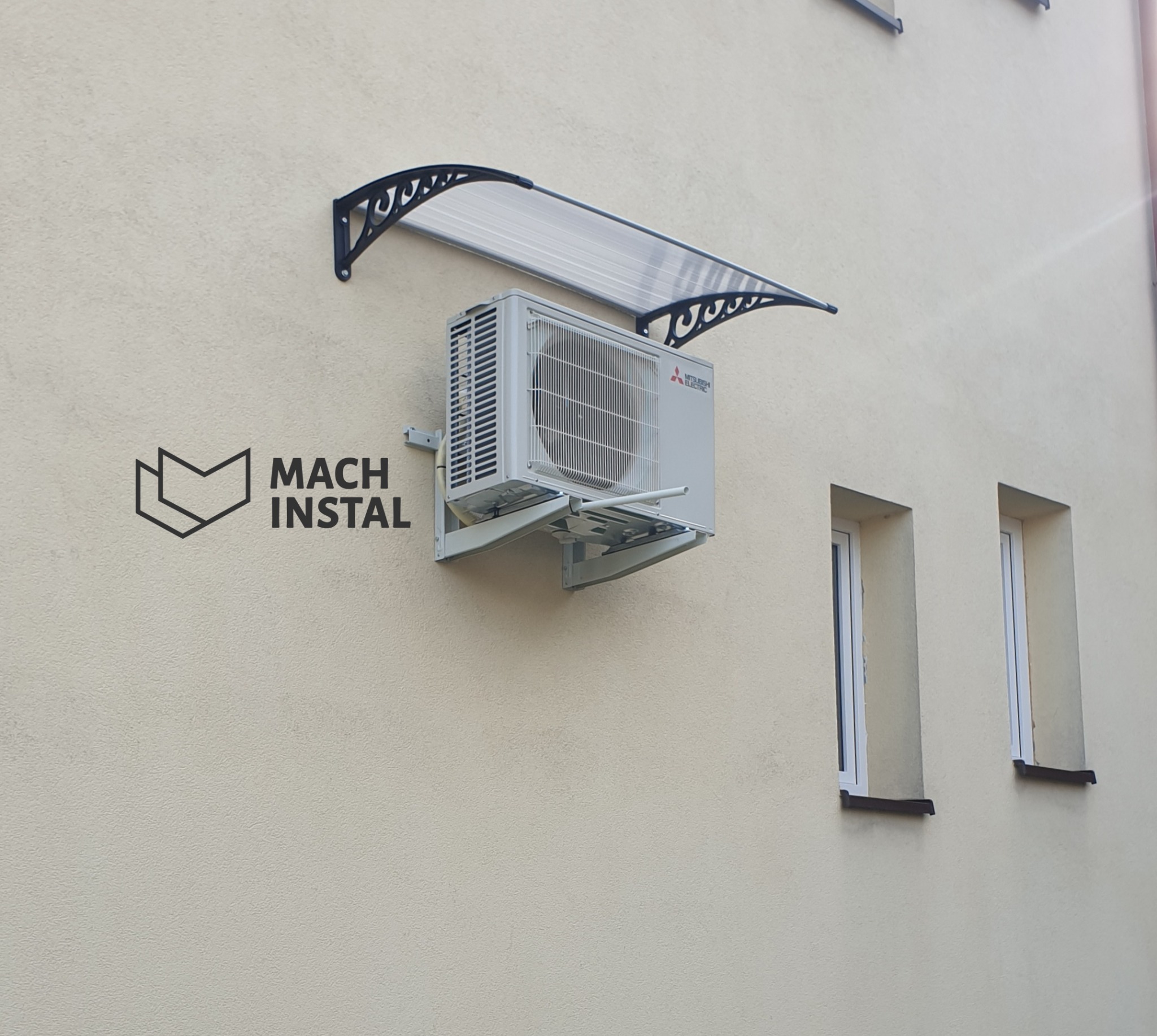 Zewnętrzna jednostka klimatyzacji Mitsubishi Electric zamontowana na elewacji budynku, osłonięta przezroczystym zadaszeniem z czarną, ozdobną ramą. Widoczne logo firmy Mach Instal na ścianie.
