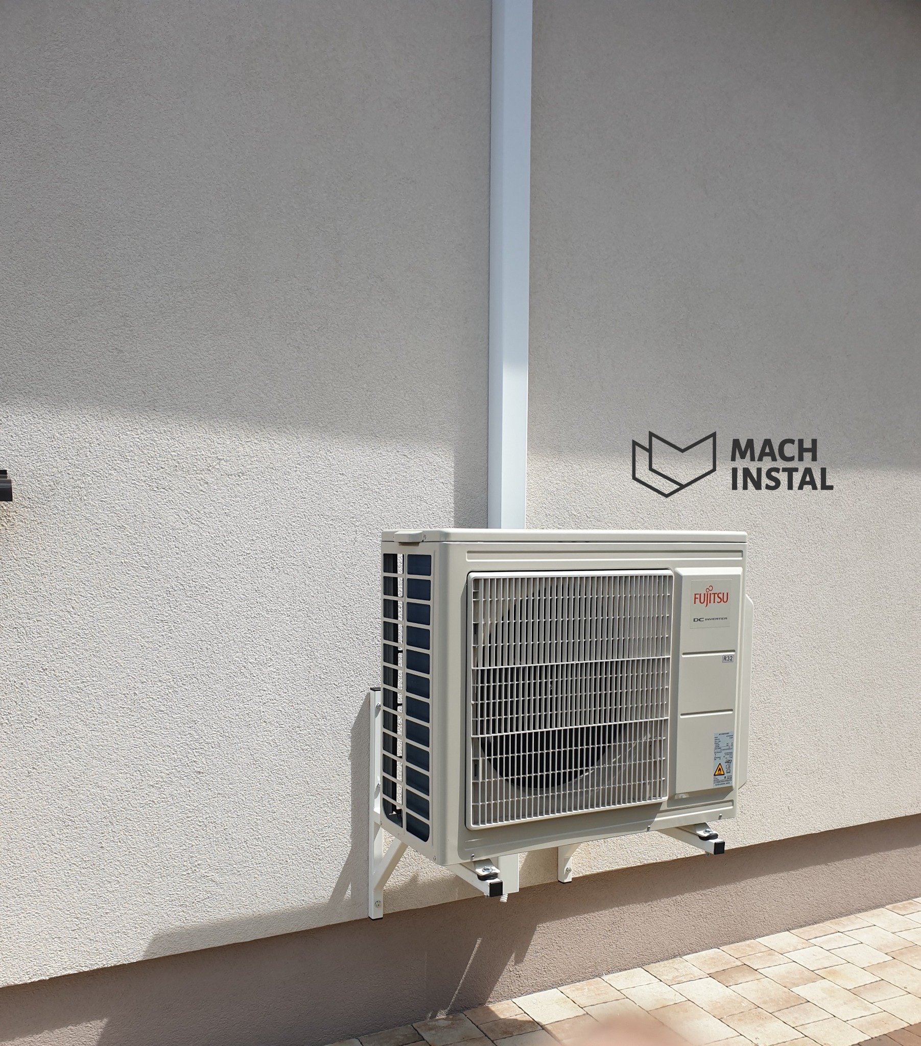 Zamontowany na elewacji zewnętrzny moduł klimatyzacji Fujitsu z widocznymi przewodami w białej maskownicy oraz logo firmy instalacyjnej Mach Instal na ścianie.