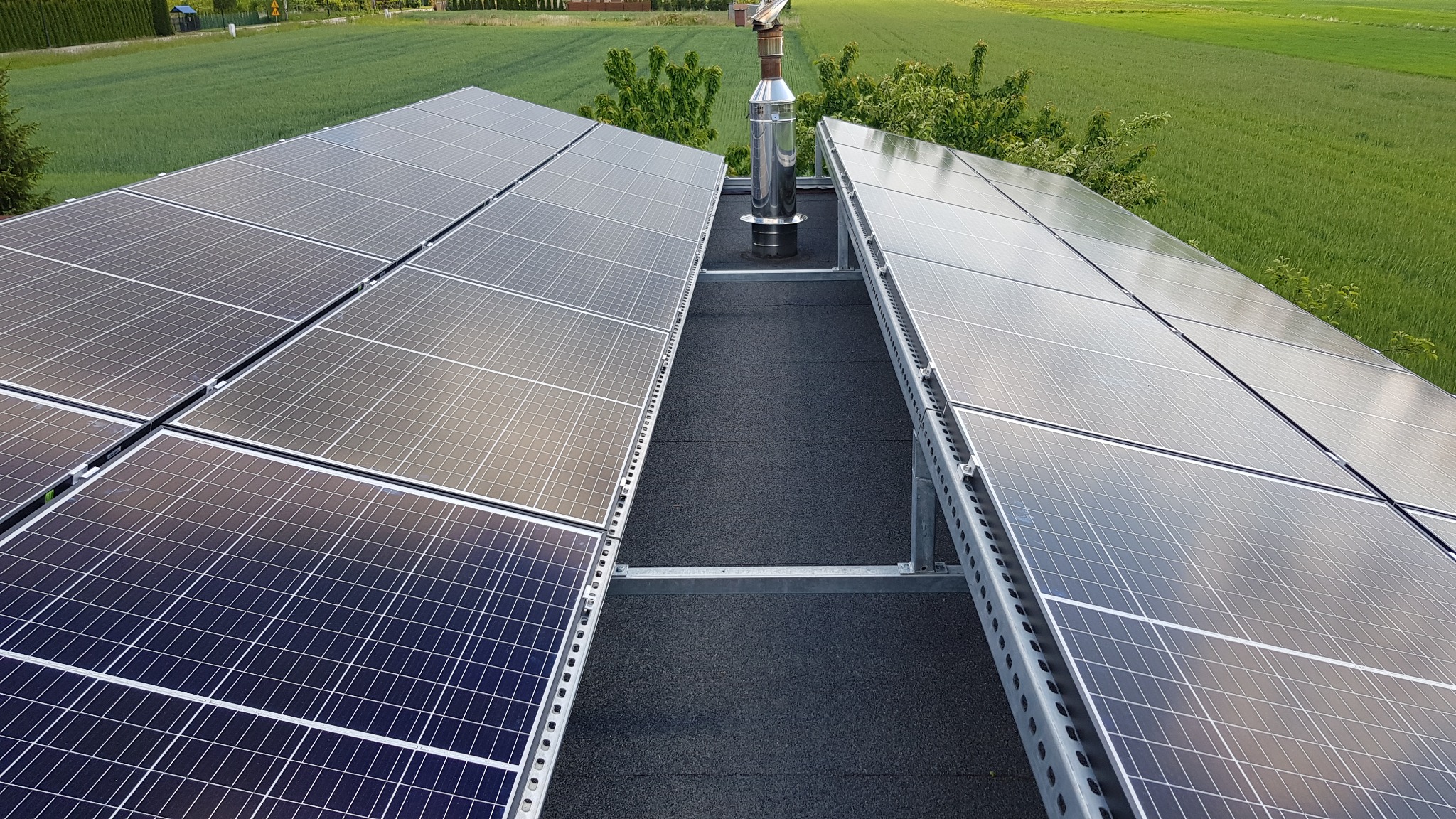 Instalacja paneli słonecznych na płaskim dachu z widocznym kominem i zielonym polem w tle.