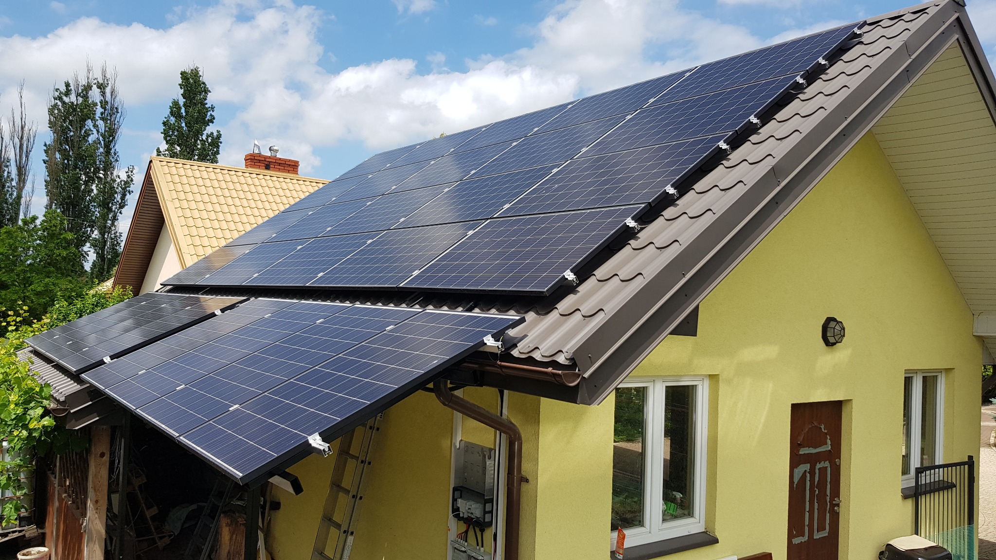 Panele słoneczne na dachu domu z żółtą elewacją, widoczna instalacja elektryczna na ścianie budynku oraz drabina oparta o ścianę.