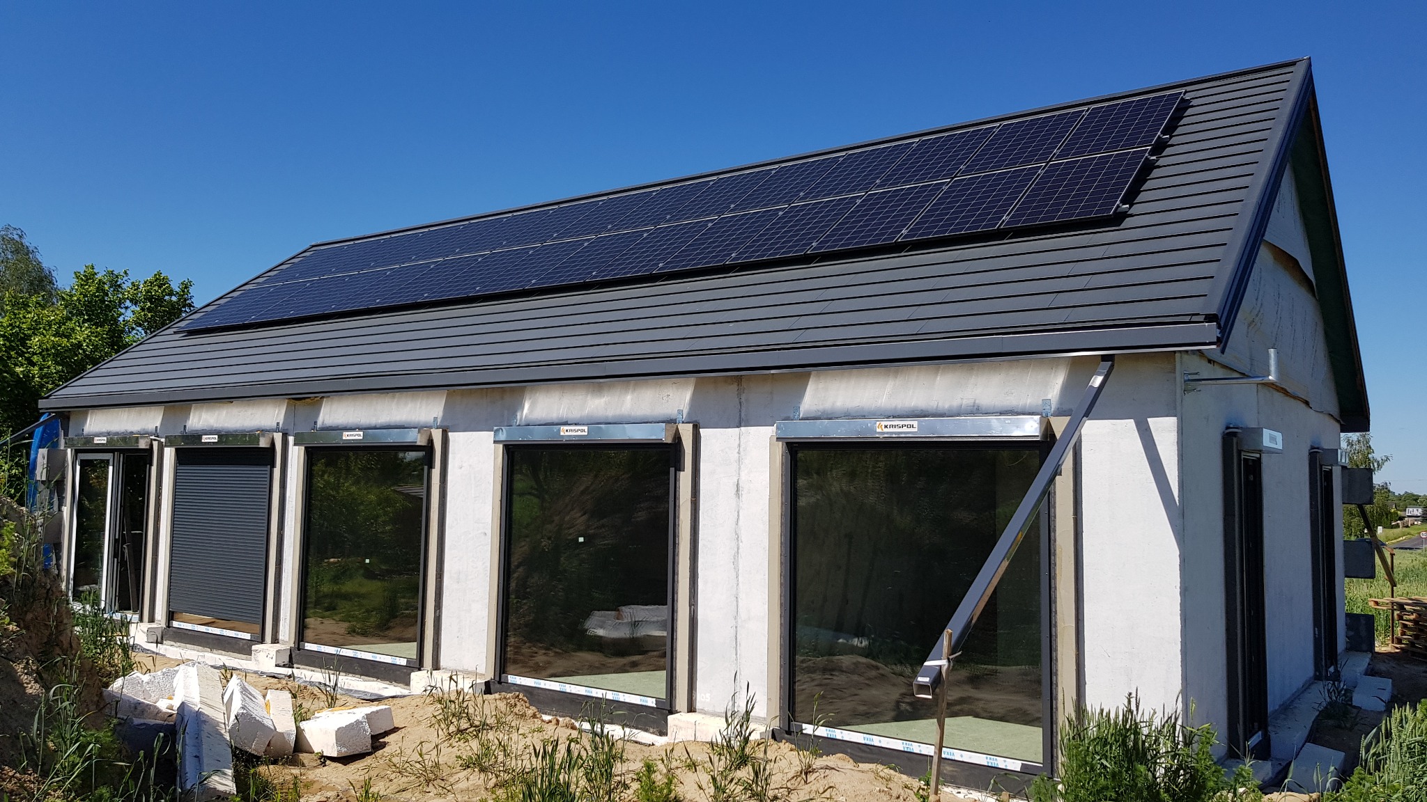 Dom jednorodzinny z zamontowanymi panelami fotowoltaicznymi na dachu pokrytym szarą dachówką, z częściowo opuszczonymi roletami zewnętrznymi marki KRISPOL.