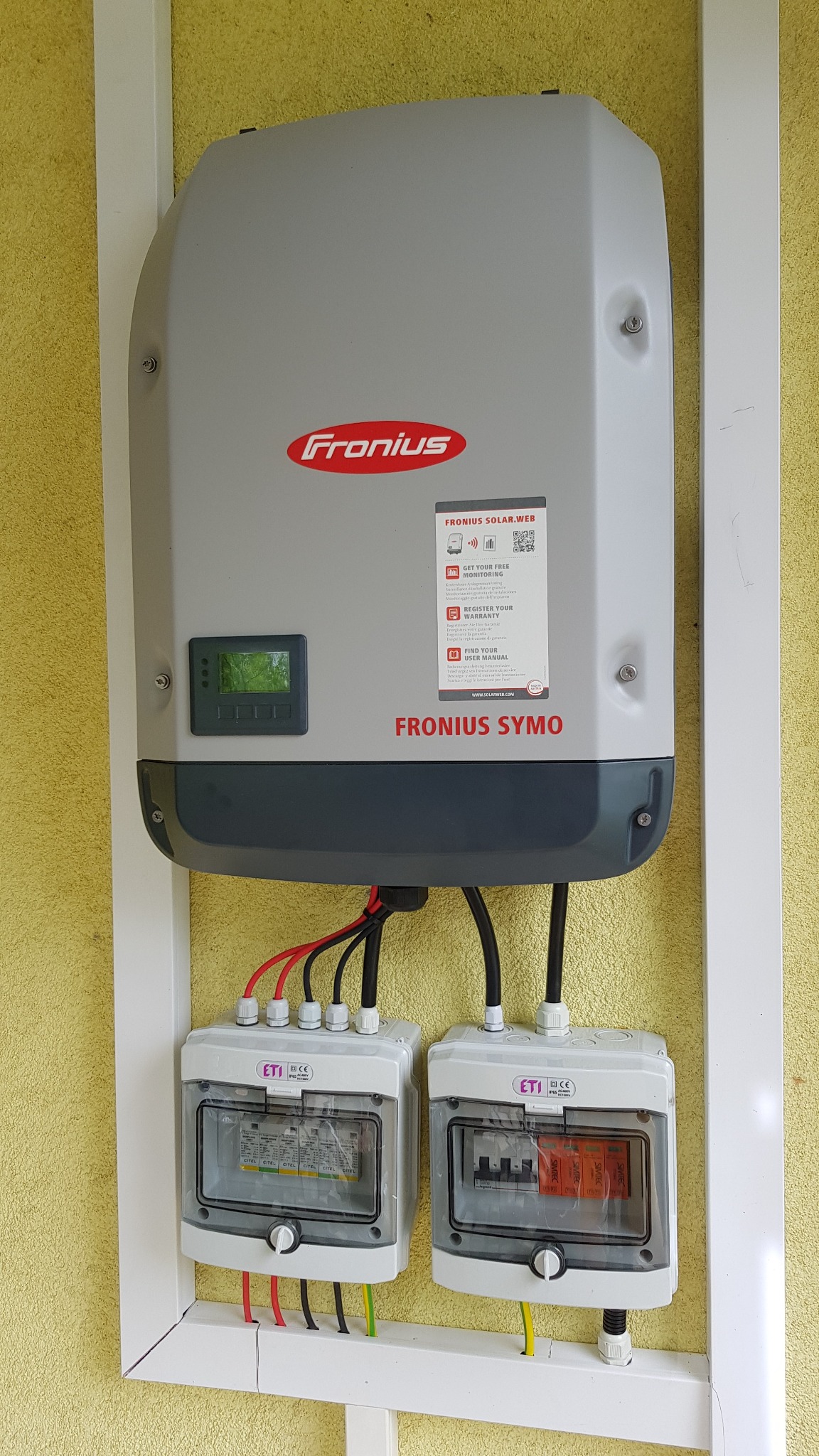 Szary inwerter Fronius Symo zamontowany na żółtej ścianie, z widocznymi dwoma białymi skrzynkami z bezpiecznikami ETI poniżej, całość w białej ramie.
