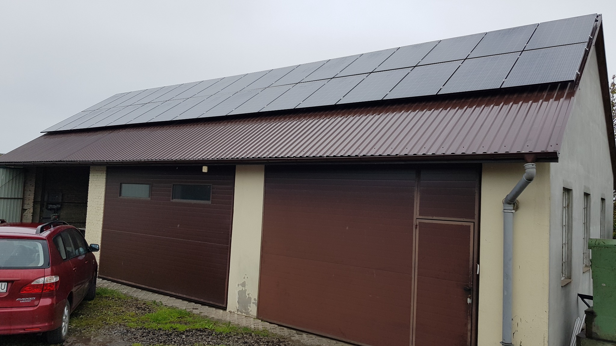 Panele słoneczne zamontowane na dachu budynku gospodarczego z brązową blachą falistą, widoczny fragment czerwonego samochodu z lewej strony.