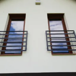 Dwa okna z zamontowanymi, prostymi, metalowymi zabezpieczeniami w formie poziomych prętów, widok z dołu na elewację budynku.