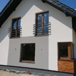 Nowoczesny dom jednorodzinny z balkonami zabezpieczonymi metalowymi barierkami o minimalistycznym designie, widoczne drewniane okna i fragment elewacji z drewnianym wykończeniem.