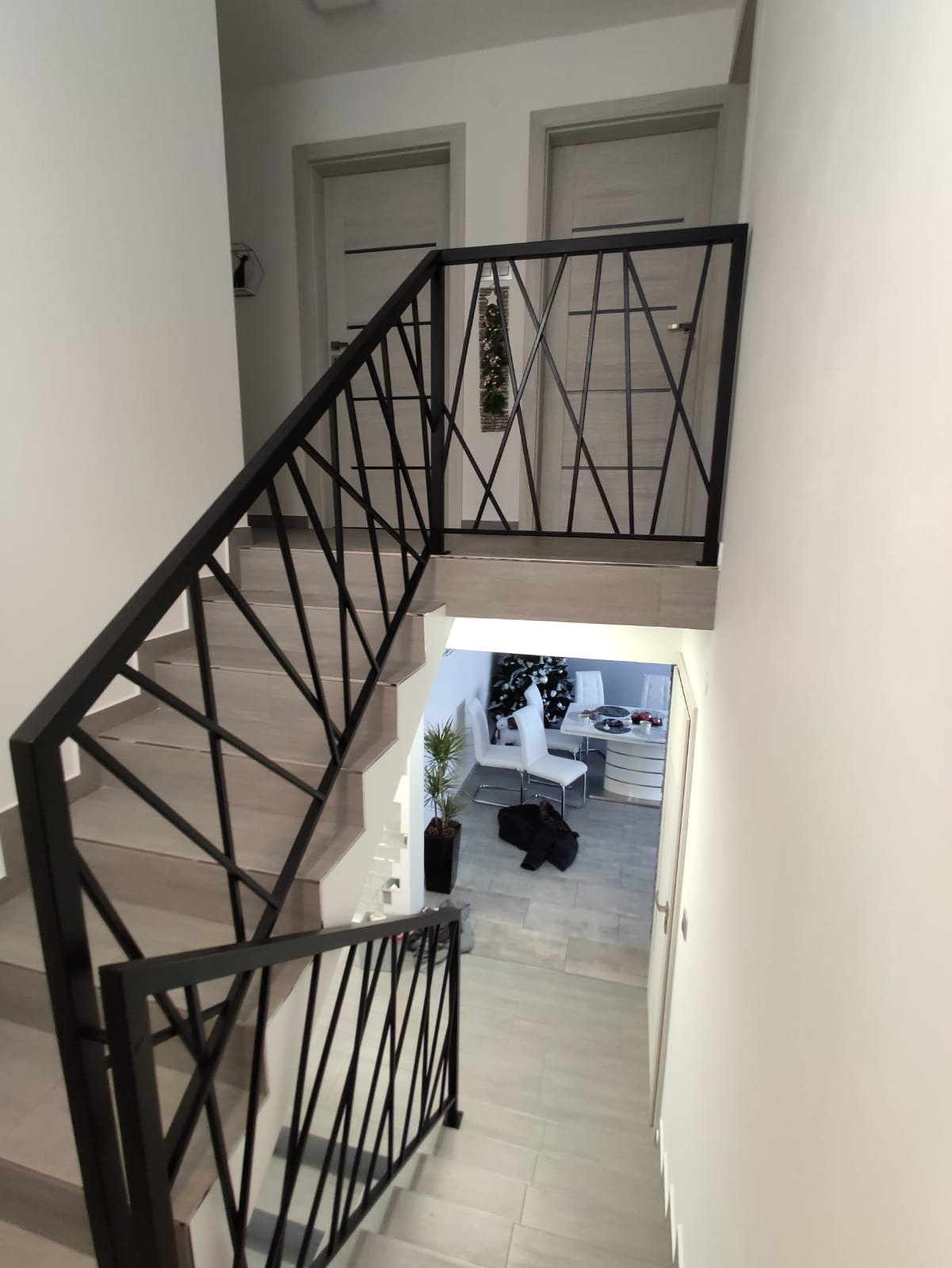 Schody wewnętrzne z czarną, geometryczną balustradą metalową, widok z góry na salon z choinką w tle.