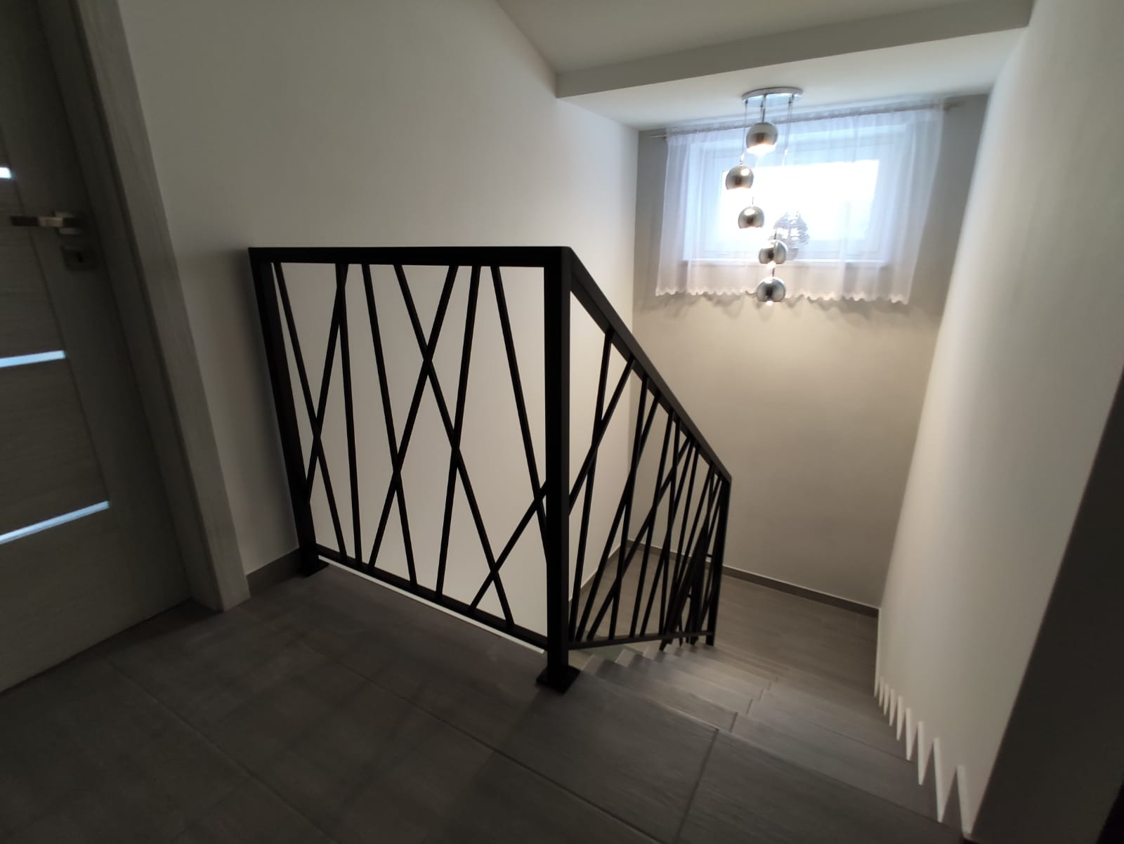 Nowoczesna balustrada schodowa z czarnymi, geometrycznymi elementami na białym tle, widok z góry na schody wyłożone szarymi płytkami, z oświetleniem w postaci wiszącej lampy z metalowymi kulami.