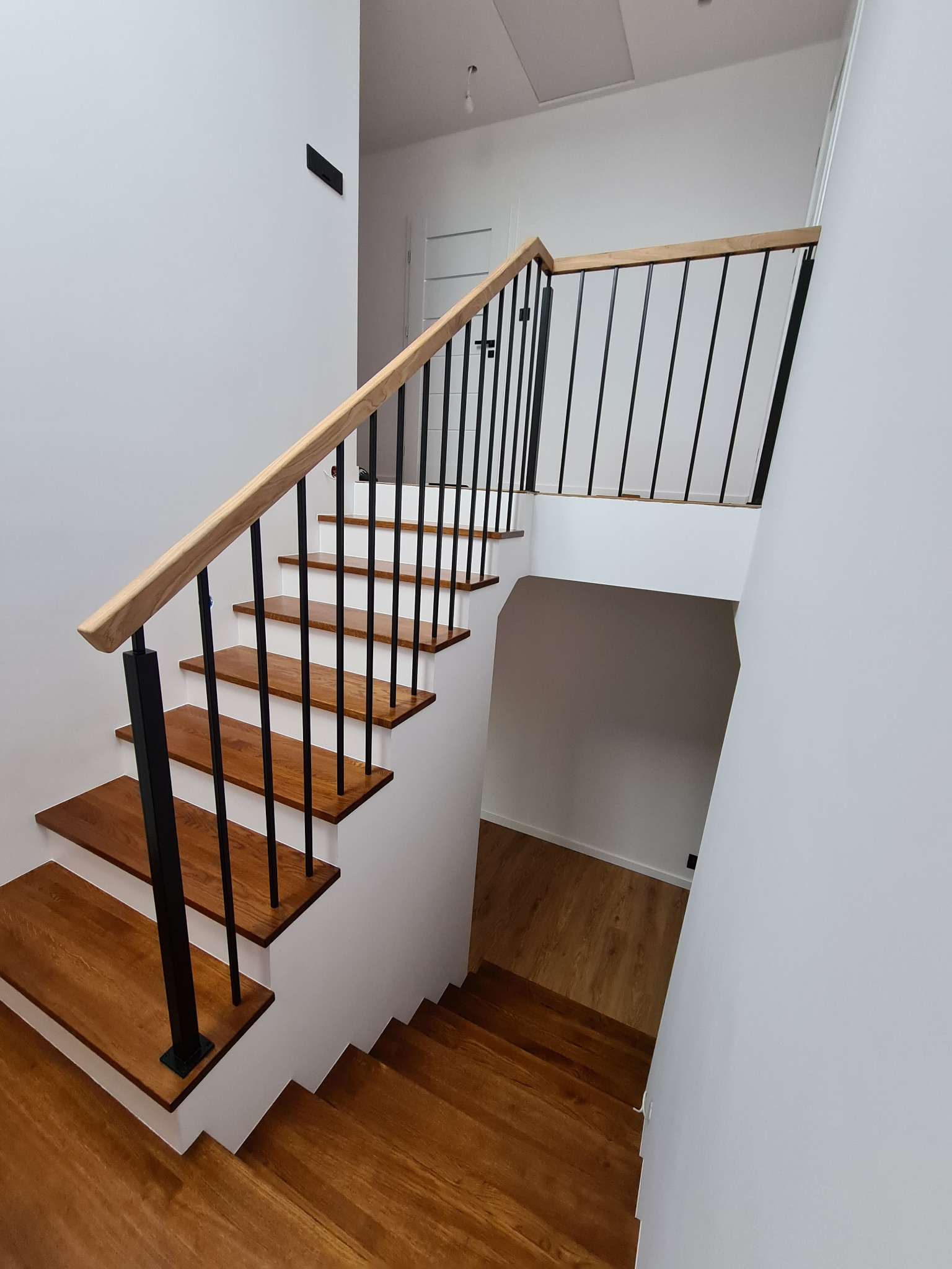 Nowoczesne schody drewniane z czarnymi metalowymi słupkami i jasną drewnianą poręczą, widok z góry na dół, minimalistyczne wnętrze z białymi ścianami.