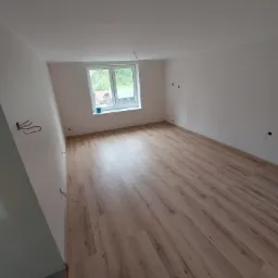 Pusty pokój po remoncie z nową podłogą laminowaną w odcieniach beżu i brązu, świeżo pomalowane ściany na biało, okno z widokiem na zieleń i wystające kable ze ściany.