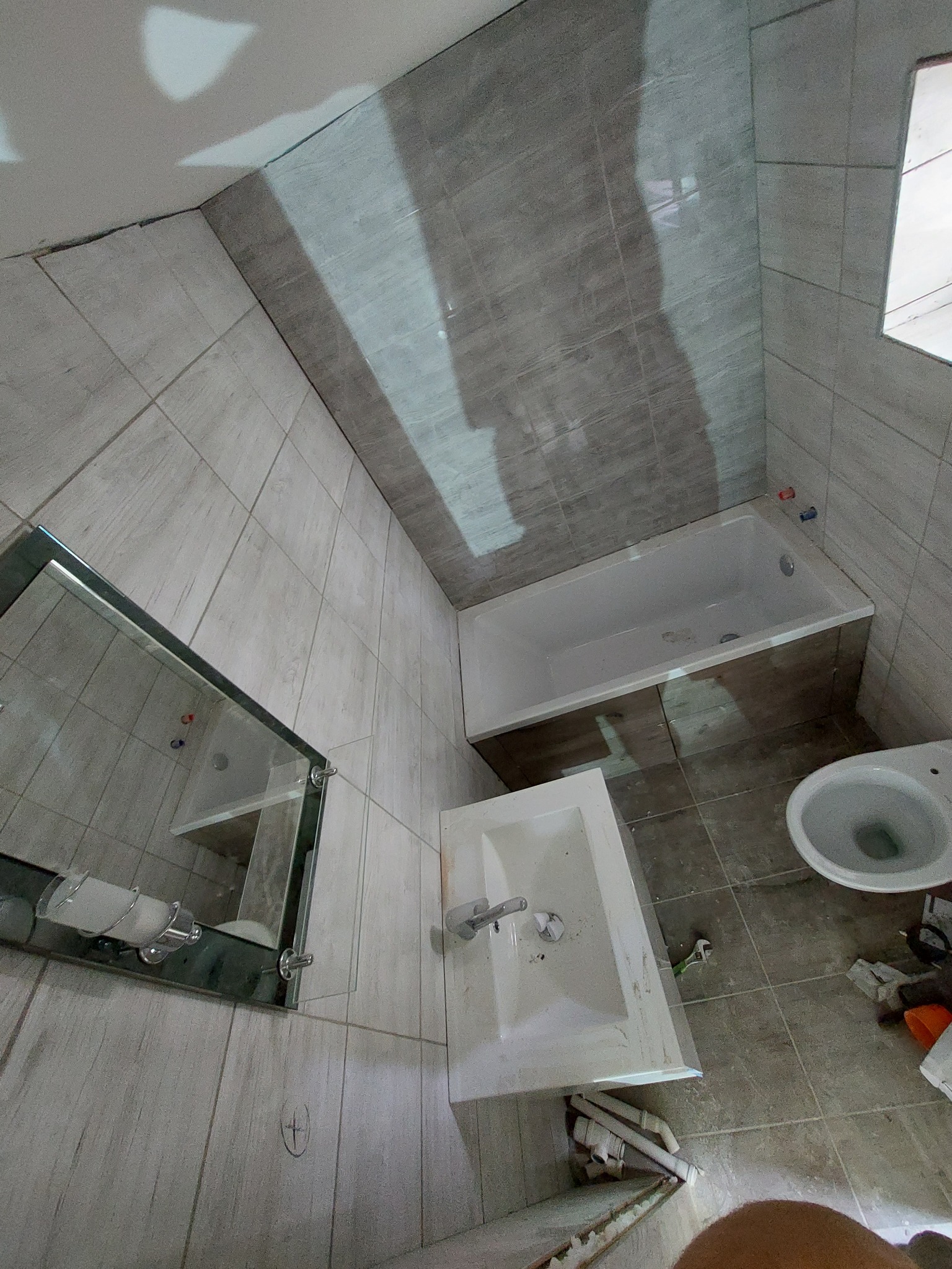 Wnętrze łazienki w trakcie prac: widoczna wanna z obudową, umywalka, toaleta, lustro z oświetleniem, szare płytki na ścianach i podłodze; na podłodze leżą narzędzia i pozostałości po pracach.