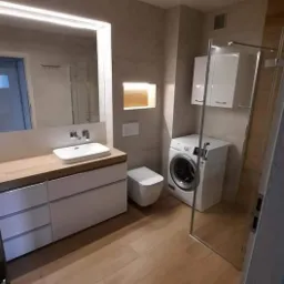 Nowoczesna łazienka z drewnianą podłogą, umywalką na szafce z szufladami, toaletą, pralką i kabiną prysznicową ze szklanymi drzwiami.