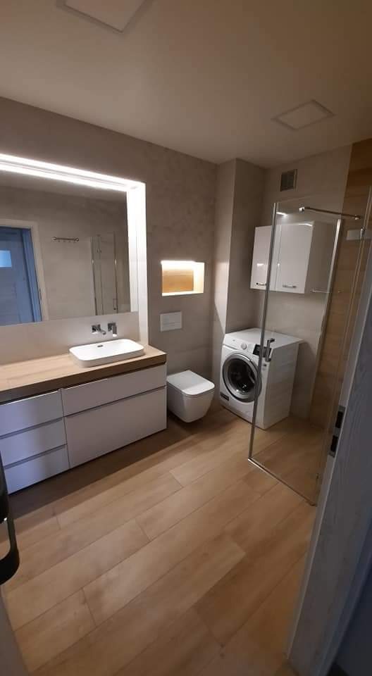 Nowoczesna łazienka z drewnianą podłogą, umywalką na szafce z szufladami, toaletą, pralką i kabiną prysznicową ze szklanymi drzwiami.