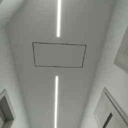 Biały sufit z dwiema podłużnymi lampami LED i otworem rewizyjnym, widok z dołu w korytarzu z drzwiami po bokach.