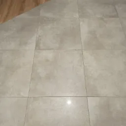 Ułożona podłoga z szarych, kwadratowych płytek ceramicznych, widoczny fragment drewnianej podłogi i biała listwa przypodłogowa