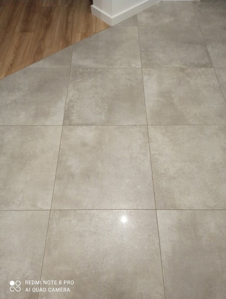 Ułożona podłoga z szarych, kwadratowych płytek ceramicznych, widoczny fragment drewnianej podłogi i biała listwa przypodłogowa