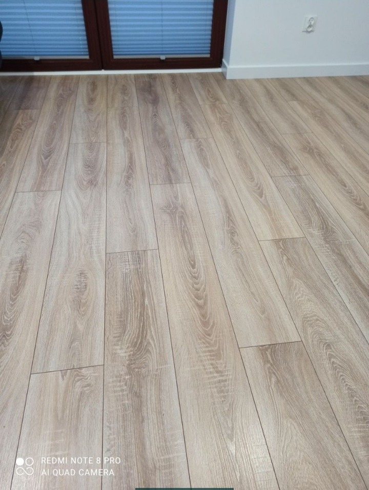 Jasne panele podłogowe ułożone wzdłuż okna z żaluzjami poziomymi, widoczne łączenia między panelami, biała listwa przypodłogowa.