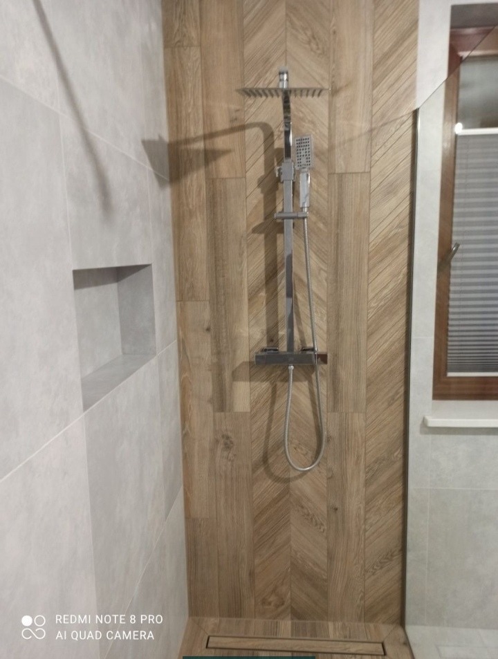 Nowoczesna kabina prysznicowa z drewnopodobnymi płytkami ułożonymi w jodełkę na ścianie, odpływ liniowy w podłodze, szara ściana z wnęką.