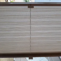 Beżowa plisa okienna z delikatnym wzorem poziomych pasków, widoczny mechanizm sterujący sznurkiem.