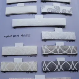 Katalog próbek tkanin do rolet i żaluzji z różnymi wzorami, ułożonych na szarym tle, z widocznymi numerami identyfikacyjnymi i informacjami o składzie.