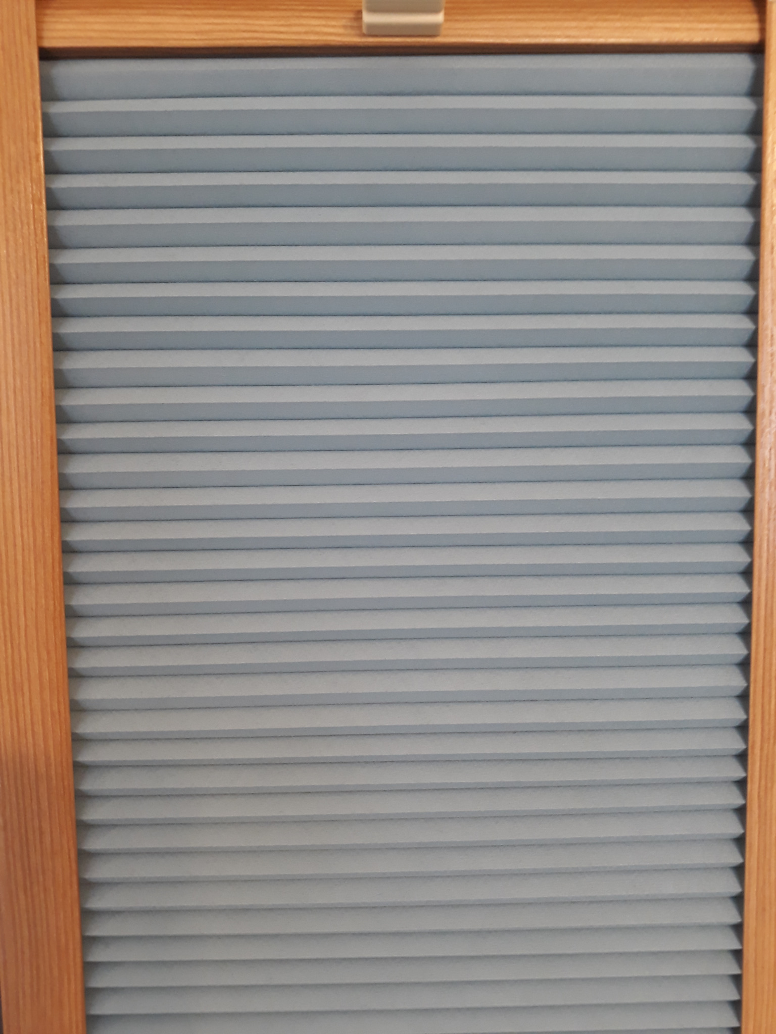 Jasnoszare, plisowane osłony okienne w drewnianej ramie, widoczne z bliska.