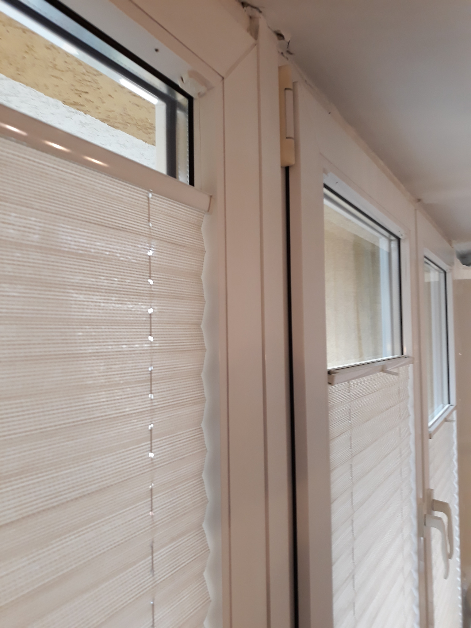 Zamknięte, kremowe plisy okienne zamontowane w białej ramie okiennej, widoczne z bliska, z delikatnym efektem rozproszenia światła.