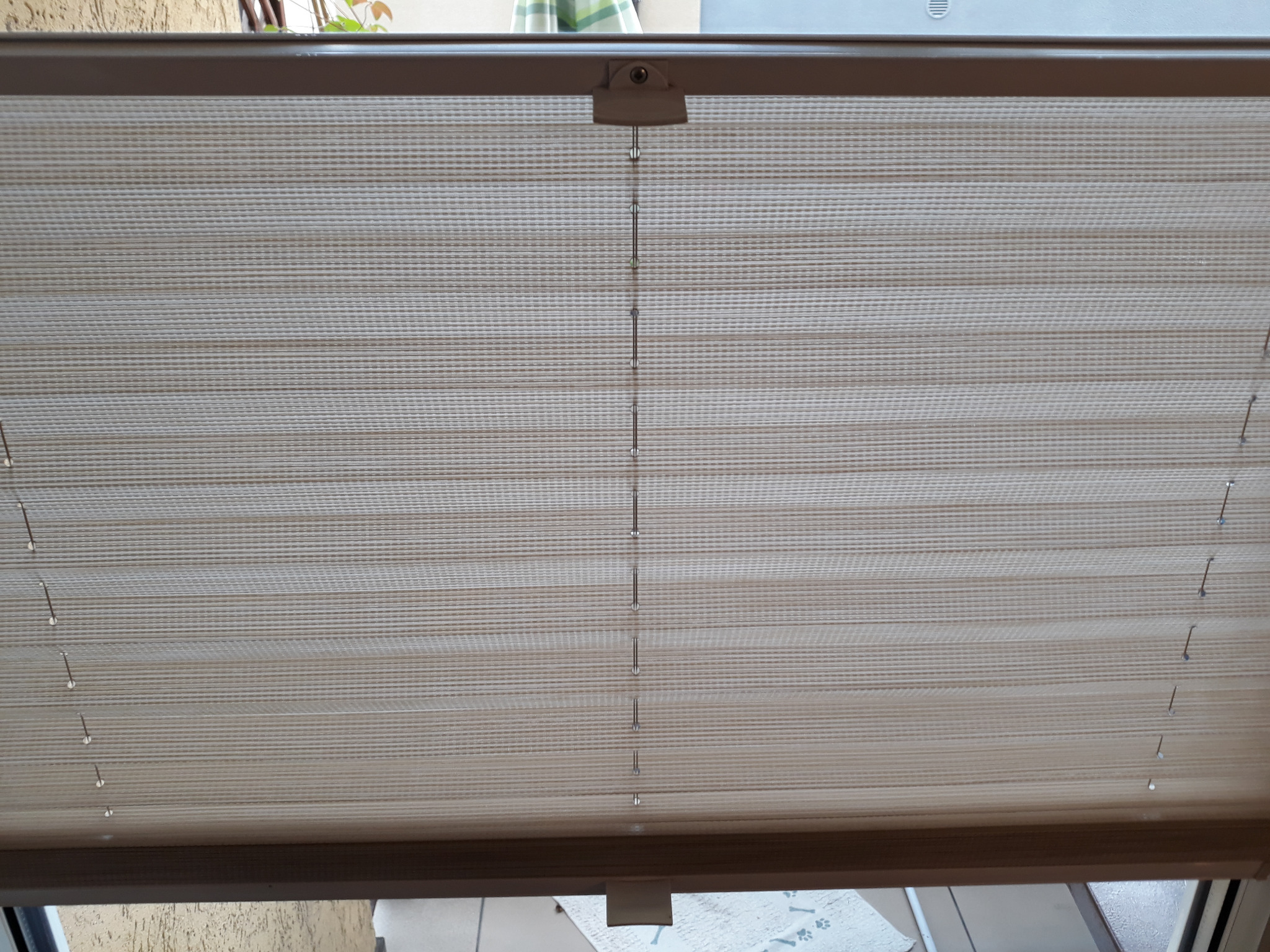 Beżowa plisa okienna z delikatnym wzorem poziomych pasków, widoczny mechanizm sterujący sznurkiem.